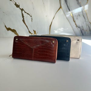 May include: Three leather wallets in a row, each with a different colour and texture. The first is a reddish-brown with a crocodile pattern, the second is black, and the third is off-white. All have a zip closure.