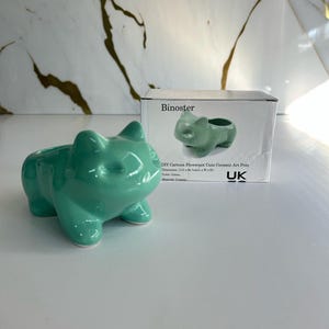 May include: A light green ceramic cat planter with a small hole in the top for planting. The planter is sitting on a white surface. The text on the box reads "DIY Cartoon Flowerpot Cute Ceramic Art Pots, Dimensions: 11.0 x 11.0 x 8.0 cm (L x W x H), Colour: Green, Material: Ceramic, UK."