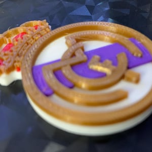 Könnte beinhalten: Eine 3D-gedruckte Medaille mit goldfarbenem Rand und Akzenten. Die Medaille hat eine weiße Mitte mit einem lila Streifen und goldfarbenen Symbolen. Ein kleines, mehrfarbiges Objekt ist seitlich angebracht.