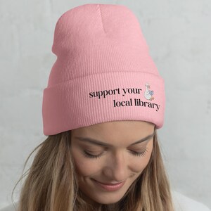 May include: A light pink beanie with the text "support your local library" in black. A cartoon goose wearing a librarian hat and holding a book is printed next to the text. The beanie is made of a soft, knitted material.