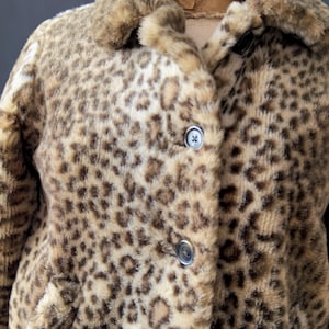 Puede incluir: Un abrigo vintage de piel sintética con estampado de leopardo. El abrigo tiene cuello con solapas, tres botones y dos bolsillos. El abrigo es de color beige, marrón y crema.