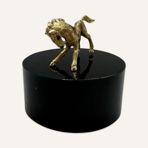 May include: A gold-colored metal figurine of a horse standing on a black circular base. The horse is in a rearing position with its head turned to the side.
