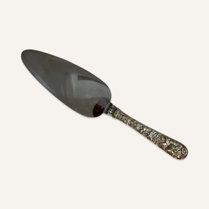 May include: A silver cake server with a wide, oval blade and a decorative handle. The handle features an ornate floral design. The server is made of a shiny, reflective metal, likely stainless steel or silver.