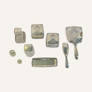 May include: A silver vanity set with a hand mirror, a tray, a small bottle, a comb, and four small boxes. The set is decorated with an ornate floral design.