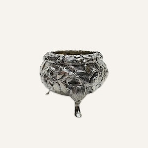 May include: A silver salt cellar with an ornate design of leaves and flowers. The cellar has three legs and is made of polished silver.