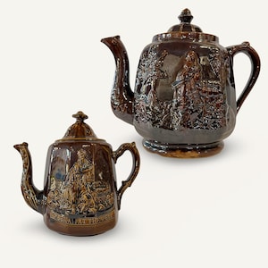 May include: Two brown ceramic teapots with detailed relief designs. One teapot is larger, and the other is smaller, both featuring a spout, handle, and lid. The smaller teapot has the text "REBEKAH AT THE WELL".