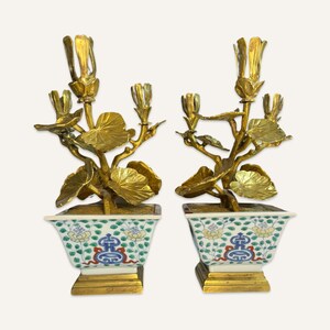 May include: A pair of gold-toned metal candle holders in the shape of stylized plants with leaves and flowers. The candle holders are in white porcelain pots with blue and green floral designs.