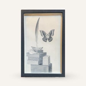 May include: Framed black and white artwork featuring a stack of wooden blocks, a feather quill, and a butterfly with a clock face. The artwork is encased in a black frame, creating a classic and elegant aesthetic.