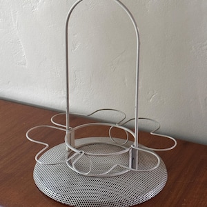 May include: A white metal wine glass holder in a flower shape. It features a circular, perforated base, a central ring, and six curved wire holders for glasses. A curved handle extends upwards from the centre.