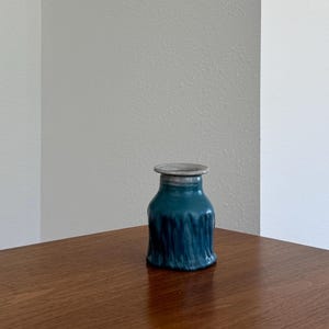May include: A small ceramic vase with a deep teal, vertically textured body and a smooth, light grey rim. The vessel features a wide, flat-topped opening that tapers into a rounded shoulder, transitioning into a slightly flared, ridged base.