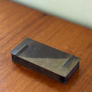 May include: A rectangular, antique metal box with a dark, aged patina. The box has a lid with two raised, decorative rectangular accents. The box is set on a wooden surface.