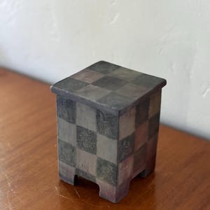 May include: A small, square, decorative box with a checkerboard pattern in shades of gray, brown, and green. The box has a slightly raised lid and rests on four small feet. The box is made of a ceramic material.