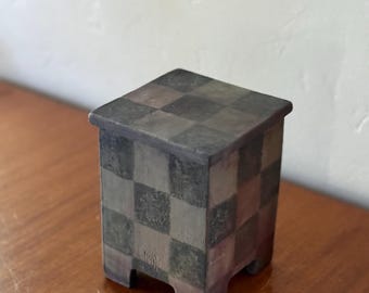 Op de afbeelding: Een kleine, vierkante decoratieve doos met een dambordpatroon in tinten grijs, bruin en groen. De doos heeft een iets verhoogd deksel en staat op vier kleine voetjes. De doos is gemaakt van keramiek.