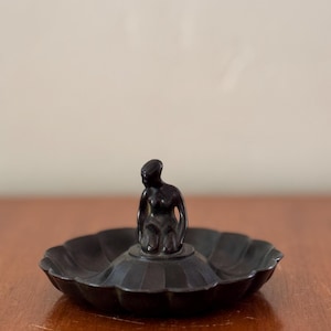 May include: A dark, antique-style ashtray featuring a central figurine of a seated woman. The ashtray has a scalloped edge and a dark finish, possibly bronze. The figurine is also dark, creating a cohesive aesthetic.