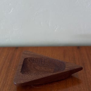 May include: A triangular wooden dish with a carved interior. The dish is a rich, dark brown color and sits on a wooden surface. The dish has a smooth, polished finish.