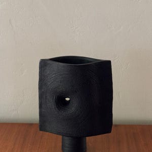 May include: A black, textured ceramic vase with a square body and a circular opening. The vase has a cylindrical base and a hole in the center of the square. The vase is displayed on a wooden surface.