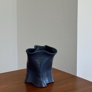 May include: A dark blue ceramic vase with a smooth, matte finish and a distinctively irregular, wavy rim. The body of the vessel features soft, vertical folds and a flared base, creating a fluid, organic shape that tapers slightly toward the center.