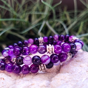 Puede incluir: Un conjunto de tres pulseras de ágata púrpura con cuentas espaciadoras plateadas. Las pulseras están apiladas una encima de la otra.