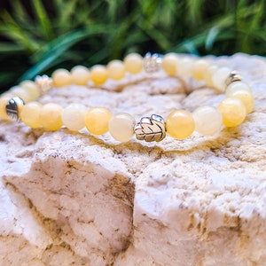 Op de afbeelding: Een gele en witte kralen armband met zilveren bladvormige bedels. De armband ligt op een witte steen.
