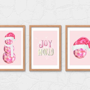 May include: Three pink and white Christmas-themed illustrations in wooden frames. The left image shows a pink disco ball snowman wearing a Santa hat and a white scarf. The middle image shows the text "JOY to the WORLD" in pink and yellow letters. The right image shows a pink disco ball with a Santa hat.