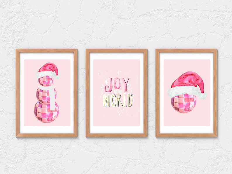 May include: Three pink and white Christmas-themed illustrations in wooden frames. The left image shows a pink disco ball snowman wearing a Santa hat and a white scarf. The middle image shows the text "JOY to the WORLD" in pink and yellow letters. The right image shows a pink disco ball with a Santa hat.