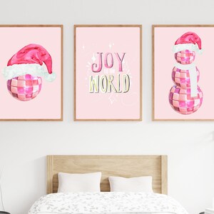 May include: Three pink and white Christmas-themed prints with a disco ball theme. The first print features a disco ball wearing a Santa hat. The second print says "Joy to the World" in a pink and gold font. The third print features a snowman made of disco balls wearing a Santa hat and a white scarf.