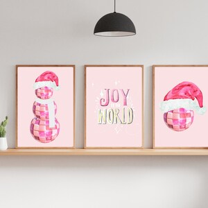 May include: Three pink and white Christmas-themed prints. The left print features a snowman made of disco balls wearing a Santa hat and a white scarf. The middle print features the text "Joy to the World" in a pink and yellow font. The right print features a disco ball wearing a Santa hat.