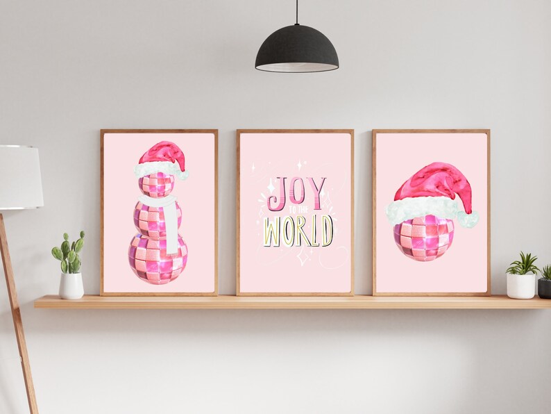 May include: Three pink and white Christmas-themed prints. The left print features a snowman made of disco balls wearing a Santa hat and a white scarf. The middle print features the text "Joy to the World" in a pink and yellow font. The right print features a disco ball wearing a Santa hat.