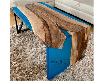 May include: A wooden coffee table with a blue epoxy resin inlay. The table has a natural wood finish and a modern design. The table is sitting on a light brown rug.