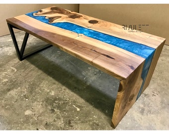 May include: A wooden coffee table with a blue epoxy resin river running through the center. The table has a black metal frame and is sitting on a concrete floor. The text "RIVULEE tables design" is visible on the table.