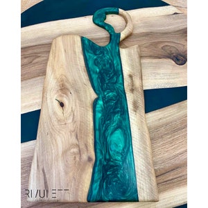 May include: A large wooden cutting board with a green epoxy resin inlay. The resin has a marbled effect and forms a unique shape in the center of the board. The board is made by Rivulet Tables Design.