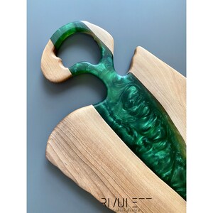 May include: A wooden cutting board with a green epoxy resin inlay. The board has a unique handle design and is perfect for serving cheese, charcuterie, or other appetizers.