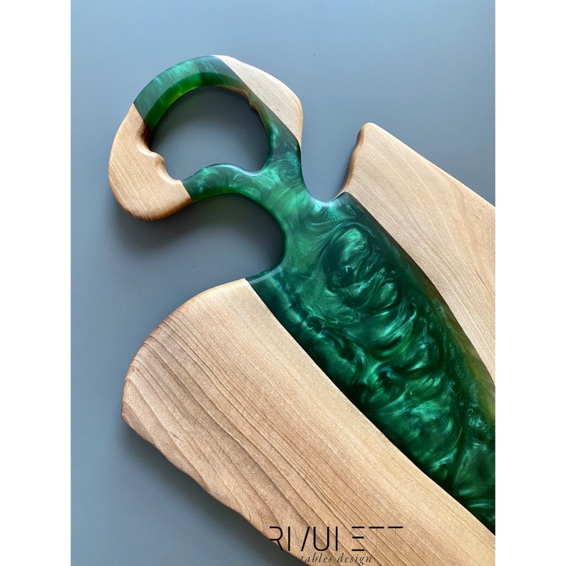 May include: A wooden cutting board with a green epoxy resin inlay. The board has a unique handle design and is perfect for serving cheese, charcuterie, or other appetizers.