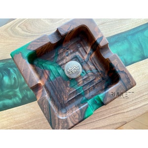 May include: A square wooden ashtray with a green and brown epoxy resin design. The ashtray has a small metal leaf design in the center.