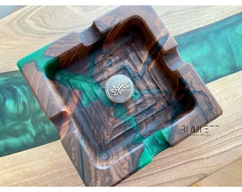 May include: A square wooden ashtray with a green and brown epoxy resin design. The ashtray has a small metal leaf design in the center.