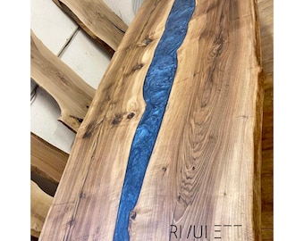 May include: A large wooden table top with a blue epoxy resin river running down the center. The table top has a natural wood grain and is finished with a clear coat. The text "RIVULET tables design" is printed on the table top.