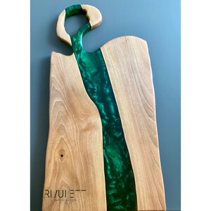 May include: A wooden cutting board with a green epoxy resin inlay. The resin has a swirling pattern and is shaped like a river. The board has a unique handle and the text "RIVULET - tables design" is printed on the bottom.