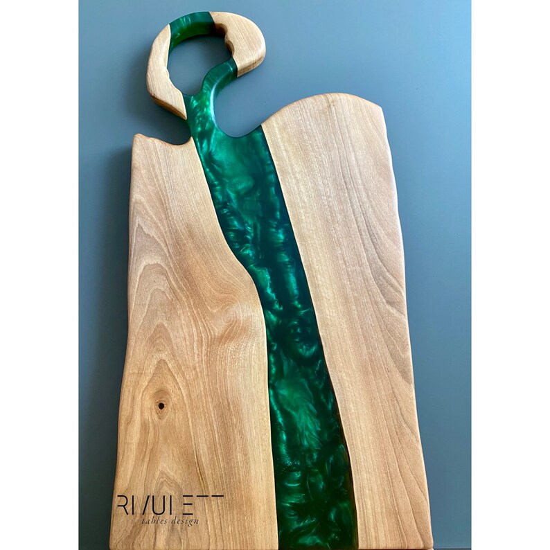 May include: A wooden cutting board with a green epoxy resin inlay. The resin has a swirling pattern and is shaped like a river. The board has a unique handle and the text "RIVULET - tables design" is printed on the bottom.