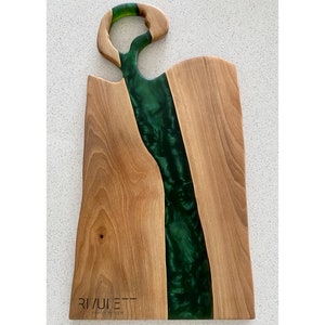 May include: A wooden cutting board with a green epoxy resin river design. The board has a unique cutout handle and the text "RIVULETT Tables Design" is printed on the bottom.