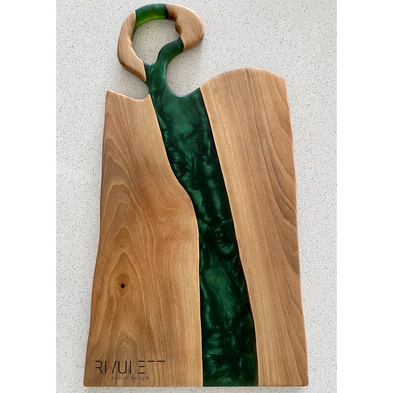 May include: A wooden cutting board with a green epoxy resin river design. The board has a unique cutout handle and the text "RIVULETT Tables Design" is printed on the bottom.