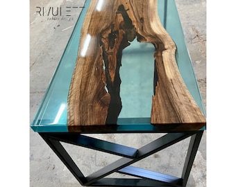 May include: A wooden table top with a blue resin river running through it. The table has a clear glass top and a black metal base with a geometric design. The table is labelled "RIVUIET tables design".