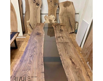 Puede incluir: Una mesa larga de madera con un acabado de madera natural y un río de resina epoxi transparente que recorre el centro. La mesa está hecha de una sola losa de madera y tiene un diseño de borde vivo. La mesa está sentada sobre un suelo de madera y hay otras piezas de madera en el fondo. El texto "RIVUET - tables design" es visible en la mesa.