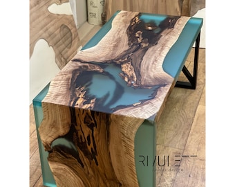 May include: A coffee table crafted from wood and epoxy resin. The resin is a light blue colour and features a wave-like pattern. The wood is a dark brown colour and has a natural grain pattern. The table is positioned on a wooden floor. The text "RIVUIER - tables design" is visible on the table.