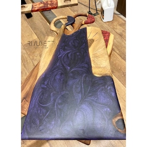 May include: A wooden serving board with a purple resin inlay. The resin has a marbled pattern. The board has a unique shape with two handles. The text "RIVULET tables design" is printed on the board.