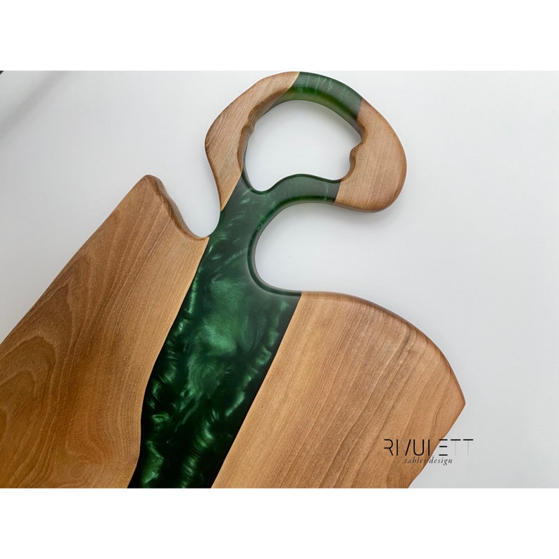 May include: A wooden serving board with a green epoxy resin inlay. The board has a unique handle with a hole in the center. The board is made by Rivulet Table Design.