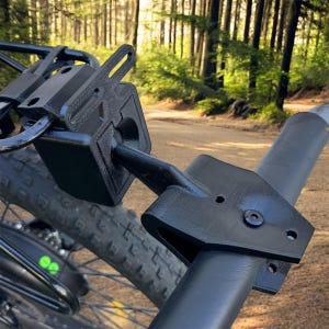 May include: Black bicycle accessory mounted on a bike frame. The accessory appears to be made of a matte black material and is designed to hold or secure something. The background shows a forest and a dirt road.