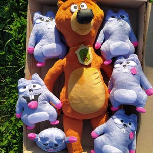 May include: A collection of plush toys featuring a large orange bear with a green and yellow emblem, surrounded by several blue hamster-like creatures with pink accents. The toys are arranged inside a cardboard box, set against a backdrop of green grass.