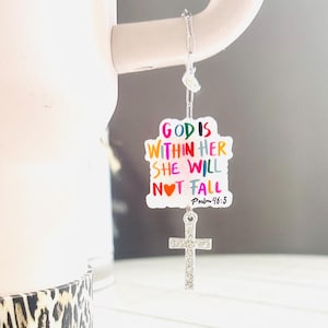May include: Two colorful charm pendants with the text "God is within her she will not fall Psalm 91:5" and a silver cross charm. The charms are hanging from a pink and white mug with a leopard print design.
