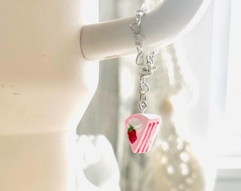 May include: A large, light pink tumbler with a leopard print sleeve. A small charm of a pink cake slice with a strawberry hangs from the handle. The tumbler sits on a white wooden surface.