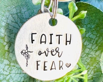Könnte beinhalten: Silberner Schlüsselanhänger mit einem runden Anhänger, auf dem "FAITH over FEAR" mit einem kleinen Herzen und einem Kreuz mit Blumen steht.